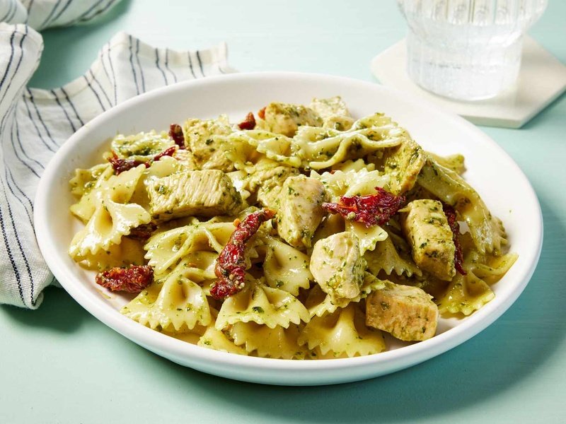 A bowl of pesto pasta with chicken next to a striped napkin and a glass of sparkling water