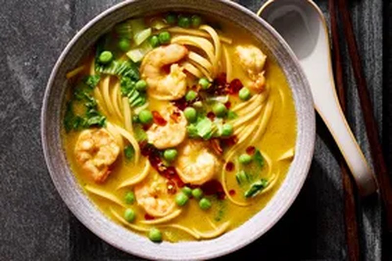 A bowl of shrimp noodle soup garnished with green vegetables and chili oil placed on a dark surface with a spoon and chopsticks nearby