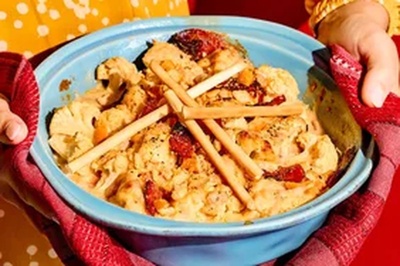A casserole dish containing cauliflower gratin with decorative breadsticks being held in hands with red kitchen towels