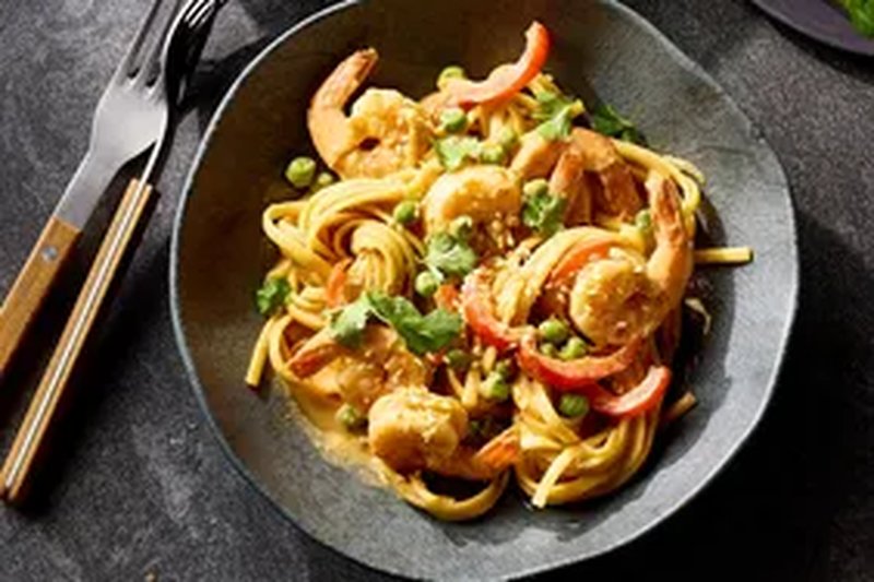 A dish of shrimp stirfry noodles topped with vegetables and herbs served in a bowl with utensils nearby