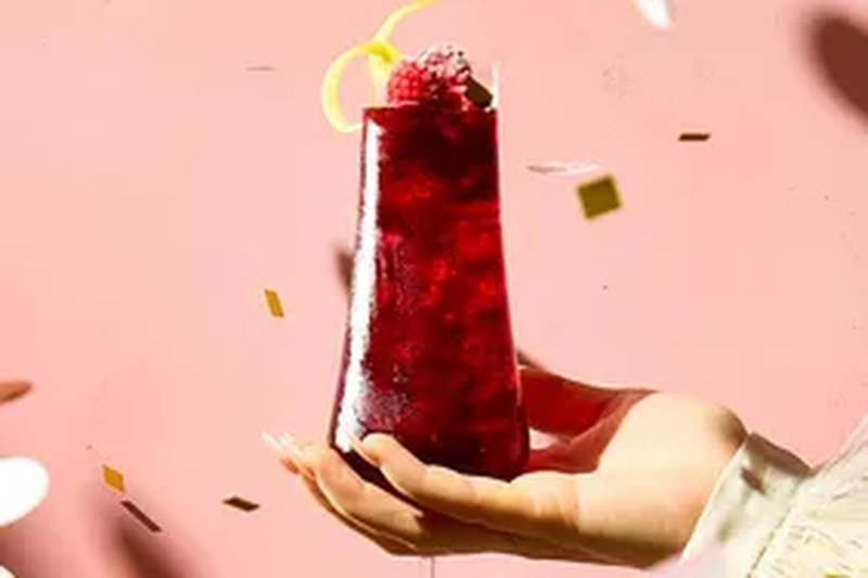 A hand holding a tall glass with a drink garnished with fruit and a lemon peel twist decorative elements in the background