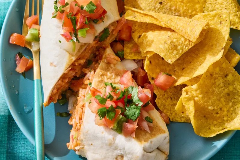 A plate with a burrito topped with diced vegetables and a serving of tortilla chips