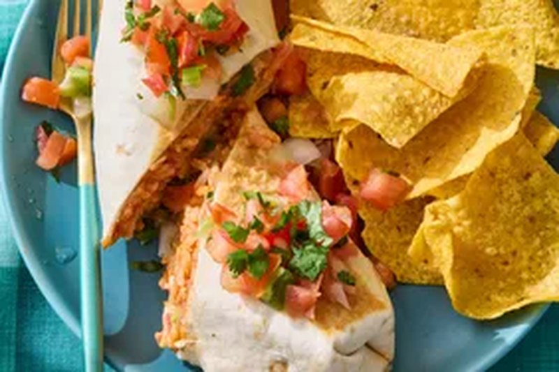 A plate with a burrito topped with diced vegetables and a serving of tortilla chips