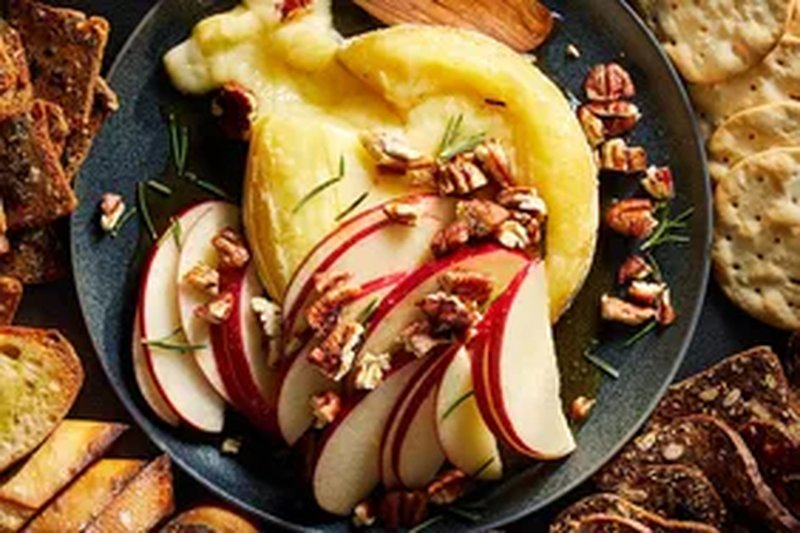 Baked brie topped with apple slices pecans and rosemary served with assorted bread and crackers on a platter
