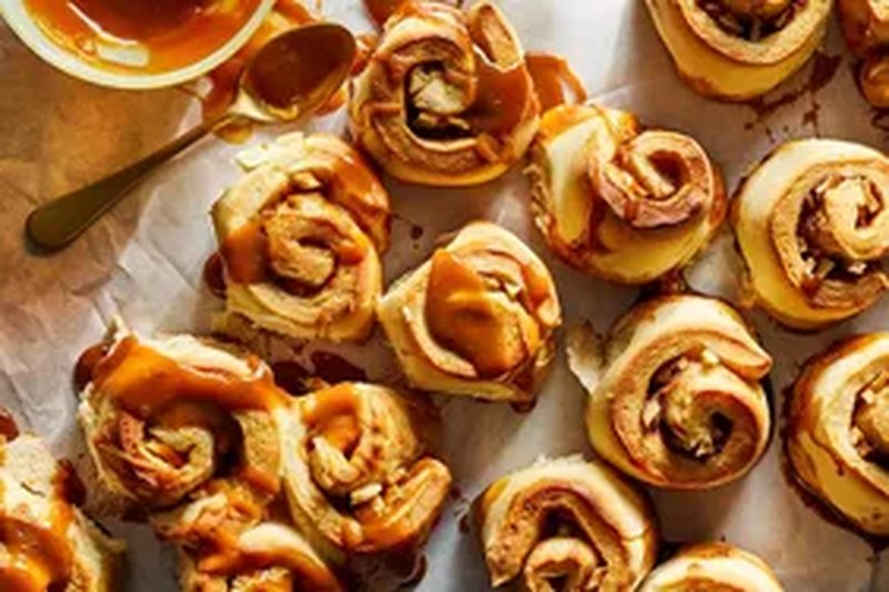 Caramel apple cinnamon rolls arranged on parchment paper with a bowl of caramel sauce and a spoon