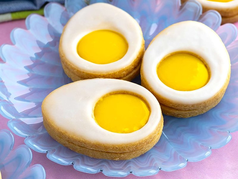 glazed egg shaped sandwich cookies linzer style with cut opening filled with lemon curd yolk on pearlescent glass blue plate
