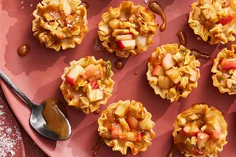 Mini tarts with brie cheese apple pieces and caramel drizzle on a plate presented alongside a spoon with caramel sauce
