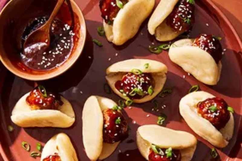 Small bao buns filled with pork and apple meatballs garnished with sesame seeds and green onions served on a platter with sauce