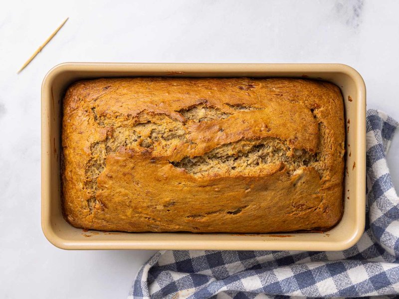 Freshly baked banana bread in a rectangular pan with a toothpick and a checkered towel beside it